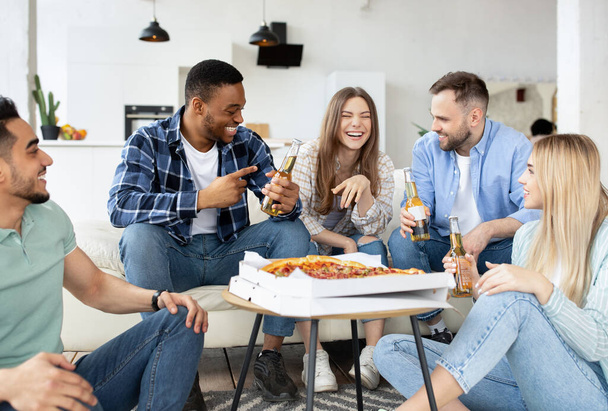 Grupa różnorodnych przyjaciół płci męskiej i żeńskiej jedzących pizzę w domu, pijących piwo, rozmawiających na imprezie studenckiej - Zdjęcie, obraz