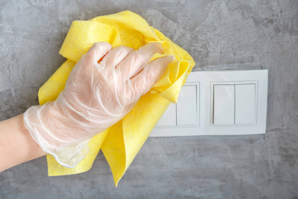 Kadın el temizleme düğmesi ve gri beton duvarda kumaş. Eldiven giymiş bir kadın, sarı bir bez parçasıyla yüzeyleri dezenfekte ediyor. Temizlikteki yeni normal Covid koronavirüsü. Yüzey dezenfeksiyonu. - Fotoğraf, Görsel