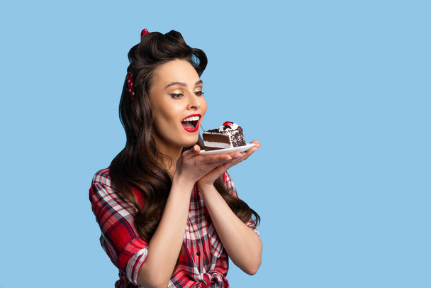 Nádherný pinup dáma v sexy retro oblečení jíst narozeninový dort na modrém pozadí studia s kopírovat prostor - Fotografie, Obrázek