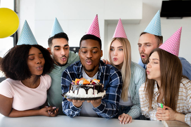 陽気な多民族の友人が誕生日ケーキの上にろうそくを吹いて、パーティーの帽子をかぶって、一緒にB日を祝う - 写真・画像
