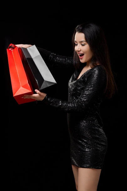 молода жінка в чорній сукні задоволена подарунком, покупками в чорно-червоних сумках. на чорному ізольованому фоні
. - Фото, зображення