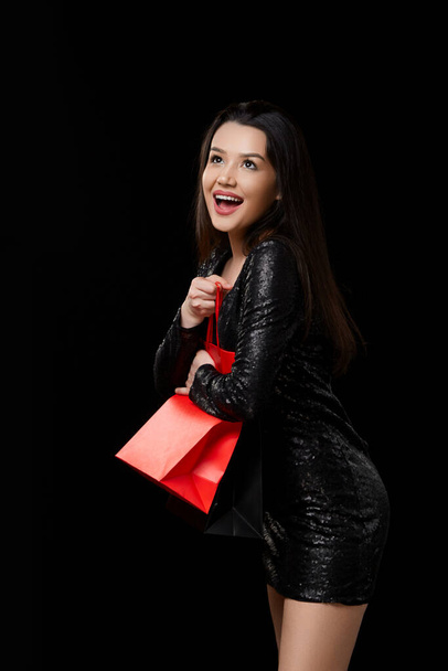 uma jovem mulher em um vestido preto está feliz com um presente, fazendo compras em sacos pretos e vermelhos. sobre um fundo isolado preto. - Foto, Imagem