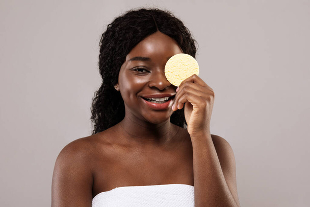 Έννοια φροντίδας δέρματος. Ελκυστική μαύρη κυρία που καλύπτει τα μάτια με καλλυντικό σφουγγάρι - Φωτογραφία, εικόνα