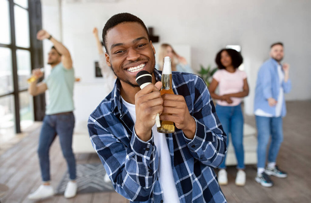 Счастливый черный парень держит бутылку пива и микрофон, исполняет песню, поет караоке на вечеринке со своими друзьями - Фото, изображение