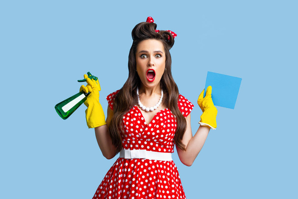 Schockierte Pinup-Frau in rotem Tupfer-Kleid mit Waschmittel und Lappen, schreit OMG auf blauem Studiohintergrund - Foto, Bild