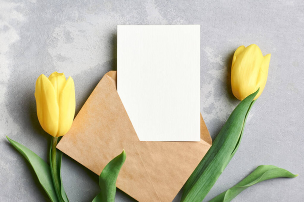 Pozdrav maketa s obálkou a žluté tulipánové květy na šedém betonovém pozadí, horní pohled, kopírovací prostor - Fotografie, Obrázek