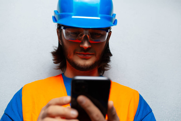 Close-up portrait of young man, construction worker engineer, looking in smartphone, wearing safety equipment on the background of grey textured wall. - Photo, Image