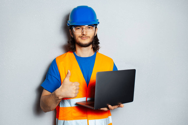若い自信家、安全装置を身に着けている建設労働者のエンジニアの肖像、ノートパソコンを保持し、灰色のテクスチャの壁の背景に親指を表示. - 写真・画像