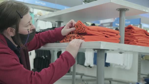 Vrouw met masker tegen virus kiest oranje shirts in kledingwinkel te kopen - Video