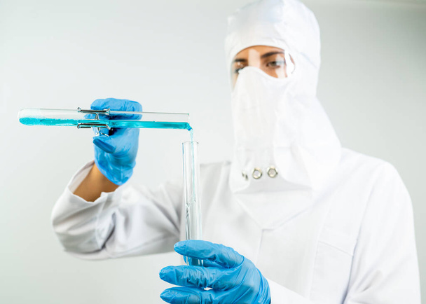 Nuori viehättävä keskittynyt naispuolinen tutkija suojalaseissa, naamiossa ja käsineissä pudottamalla sinistä nestemäistä ainetta koeputkeen pipetillä tieteellisessä kemiallisessa laboratoriossa    - Valokuva, kuva