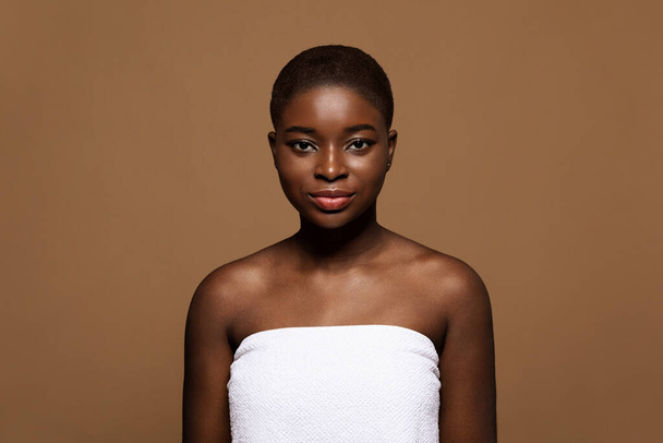 Minimalistinen meikki. Houkutteleva afrikkalainen amerikkalainen nainen yllään pyyhe seisoo ruskealla taustalla - Valokuva, kuva