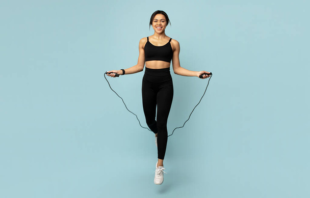 Cardio workout. Happy black woman jumping over the skipping rope, during training on blue background, panorama - Foto, immagini