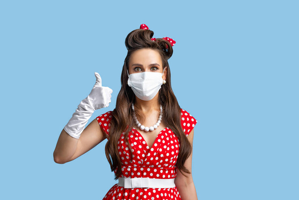 Koronavirüse karşı koruma. Yüz maskesi takan retro giysili bir kadın covid-19 'u önlemek için, baş parmak gösteriyor. - Fotoğraf, Görsel