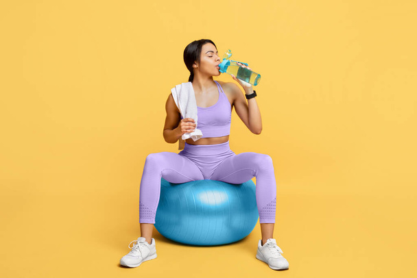 Hydratace. Arfo Fitness dívka sedí na Fitball pitné vody z láhve přes žluté pozadí. Studio Shot - Fotografie, Obrázek