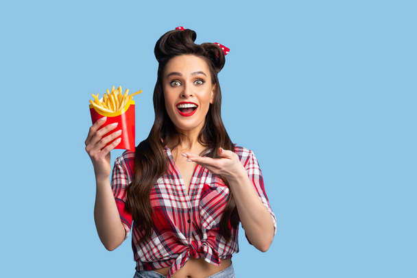 Excited young pinup woman in retro outfit holding package with yummy french fries on blue studio background - Zdjęcie, obraz