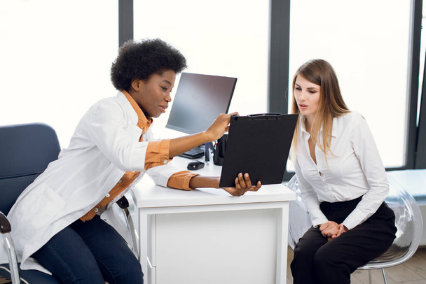 African American female doctor and pretty Caucasian woman patient Discussing the way of treatment or medical record data at modern diagnostic center. Doctor shows a clipboard to patient. - Foto, immagini