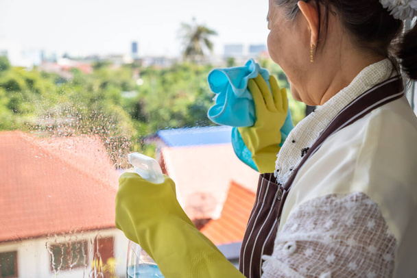 Mulher feminina sênior asiática feliz usando garrafa de spray e pano para limpar o vidro da janela - Foto, Imagem