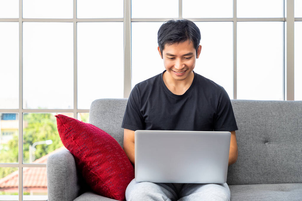 Glückliche junge asiatische Mann mit Laptop-Computer sitzen o Sofa im Wohnzimmer, arbeiten von zu Hause aus Konzept. - Foto, Bild