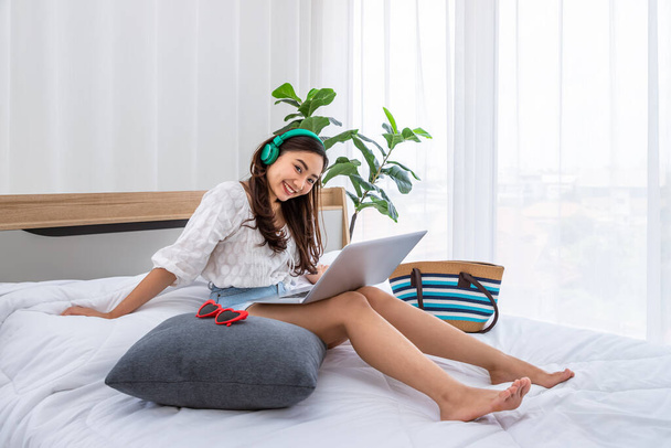 Aantrekkelijke Aziatische vrouw met koptelefoon op bed in haar slaapkamer genieten van het gebruik van laptop voor te bereiden voor de zomer vakantie reis, op zoek naar camera - Foto, afbeelding