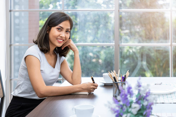 Young attractive Asian woman smiling thinking planning writing memo in notebook working from home, looking at camera - Foto, Imagem