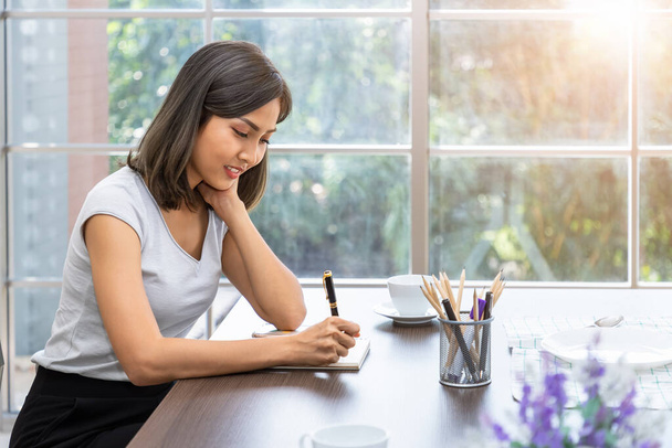 Jeune femme asiatique attrayante souriant pensée planification écriture note de service dans le carnet de travail de la maison - Photo, image