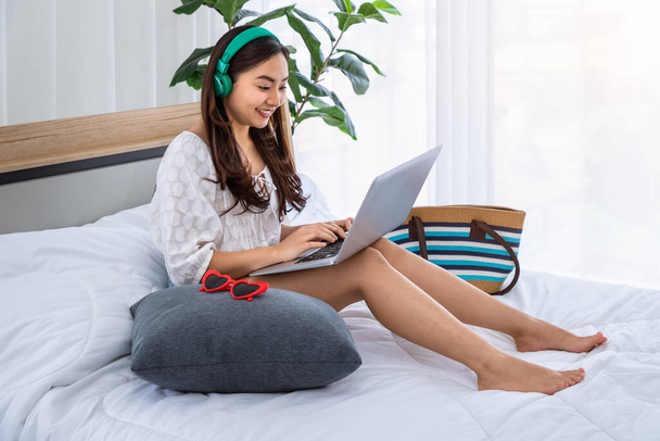 寝室にヘッドフォン付きの魅力的なアジアの女性は、夏休み旅行の準備のためにラップトップを使用してお楽しみください - 写真・画像