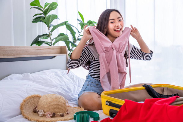 Young happy Asian woman trying clothes and packing luggage sitting on bed. Preparing for summer vacation trip. - Foto, afbeelding