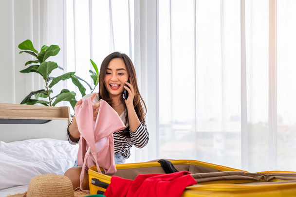 Young happy Asian woman trying clothes and packing luggage sitting on bed, talking on phone. Preparing for summer vacation trip. - 写真・画像