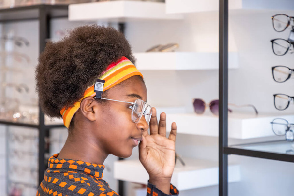 Πορτρέτο της νεαρής Αφρικής κορίτσι γυναίκα με Afro στυλ μαλλιών επιλέγοντας γυαλιά στο οπτικό κατάστημα, δοκιμάζοντας γυαλιά, κοιτάζοντας την κάμερα. Έννοια Eyecare. - Φωτογραφία, εικόνα