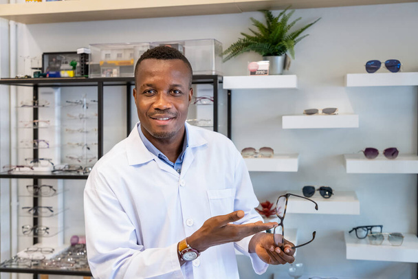 Portret van Afrikaanse opticien in optische winkel, met bril in de hand, kijkend naar de camera. Eyecare-concept. - Foto, afbeelding