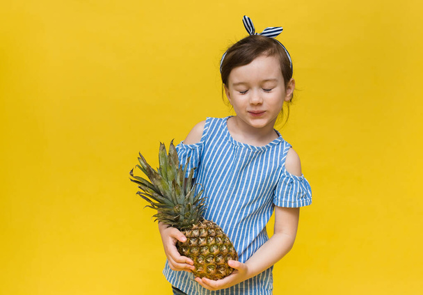 een klein meisje met een blinddoek en een gestreept T-shirt staat met gesloten ogen en houdt een ananasvrucht op een gele achtergrond met ruimte voor tekst - Foto, afbeelding