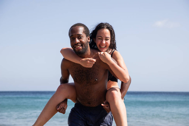Międzyrasowa para na plaży, afrykański mężczyzna podnosi białą kobietę, latem cieszą się wakacjami i dobrze się bawią - Zdjęcie, obraz