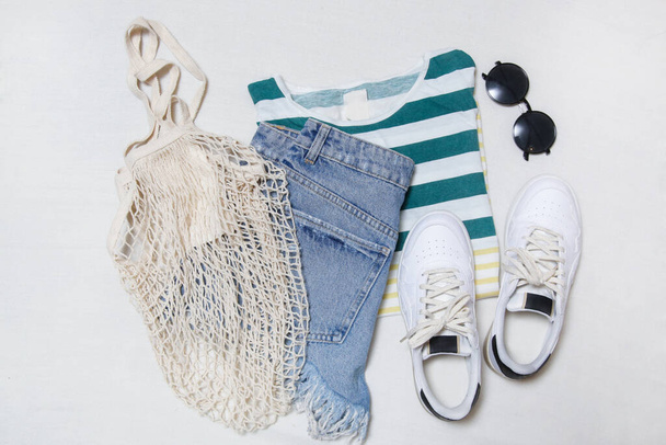 Traje de verano simple sobre fondo blanco, camisa de algodón a rayas, jeans y zapatillas blancas. Artículos de moda casual de las mujeres, vista superior. - Foto, imagen