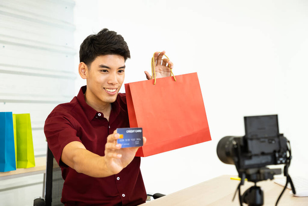 Joven asiático hombre en línea comerciante blogger utilizando la cámara de vídeo en vivo, mostrar bolsa de compras y tarjeta de crédito. Redes sociales, Influencer, compras en línea y concepto de pago. - Foto, Imagen