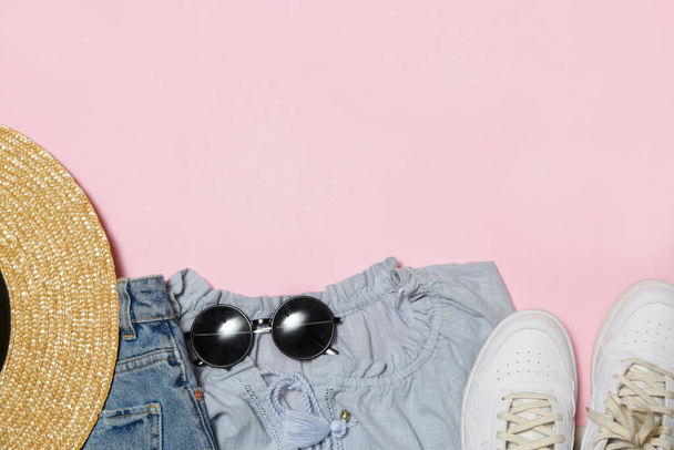 Simple summer outfit on pink background with copy space, shirt, blue jeans, straw hat, black sunglasses and white sneakers. Women's fashionable items, top view. - Photo, Image