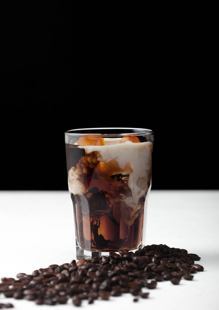 Glass of black coffee with ice cubes and fresh milk on white board with fresh coffee beans on black background. - Foto, Imagen