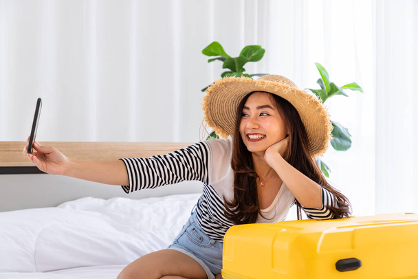 Happy Asian kobieta planuje przygotowanie podróży bagażu walizki, siedzi na łóżku w sypialni i selfie wideo połączenie - Zdjęcie, obraz