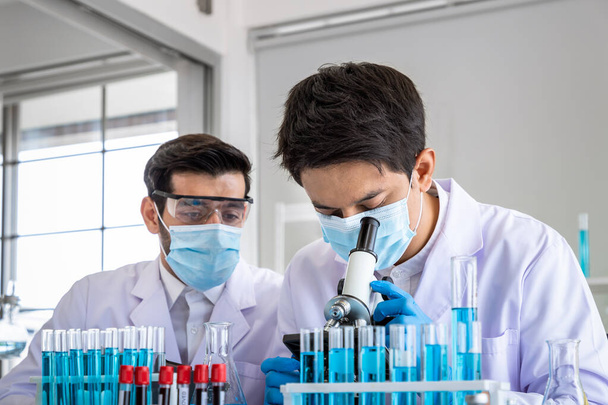 Twee mannelijke Aziatische en Arabische onderzoekers werken in het laboratorium, het uitvoeren van studie biohazard stof met wetenschappelijke apparatuur en microscoop - Foto, afbeelding