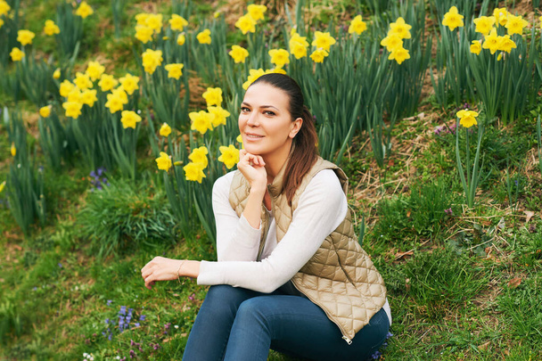 Frühlingsporträt einer jungen schönen Frau, die in einem Feld blühender Narzissen sitzt - Foto, Bild