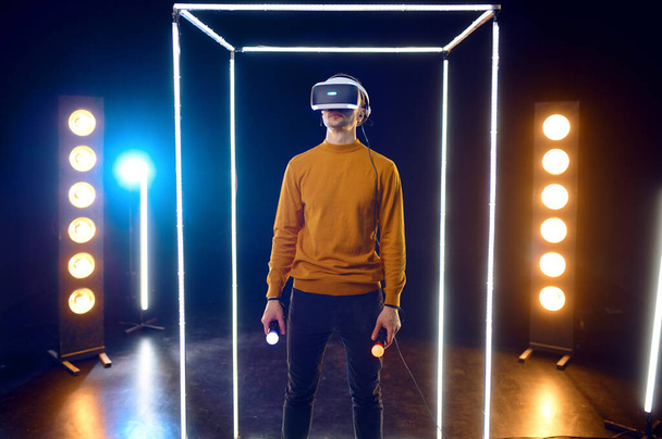 Gamer joue le jeu en utilisant un casque de réalité virtuelle et une manette de jeu en cube lumineux. Intérieur sombre du club de jeu, projecteur sur l'arrière-plan, technologie VR 3D - Photo, image