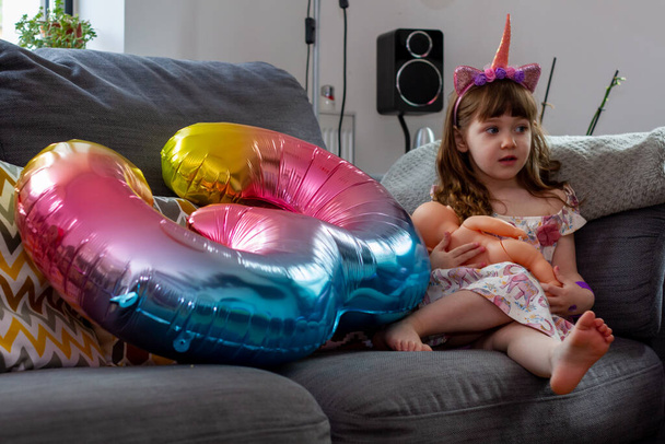 Una linda niña sentada en un sofá con un globo de colores y una muñeca - Foto, imagen