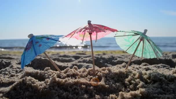 Paraguas de cóctel de papel en arena en la orilla del mar en el soleado primer día de verano - Imágenes, Vídeo