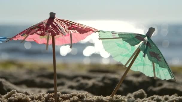 Paraguas de cóctel de papel en arena en la orilla del mar en el soleado primer día de verano - Metraje, vídeo
