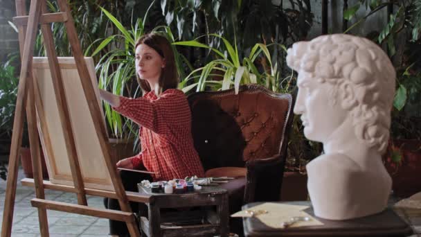 Artysta w pracowni sztuki z roślinami - młoda ładna kobieta rysująca grecką rzeźbę głowy - Materiał filmowy, wideo