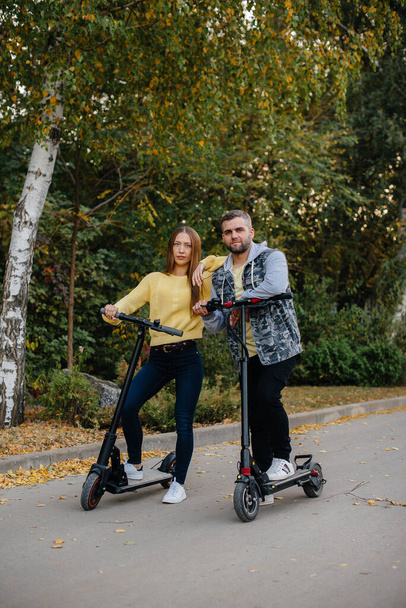 A young beautiful couple rides electric scooters in the Park on a warm autumn day. Hobbies and recreation. - Фото, изображение