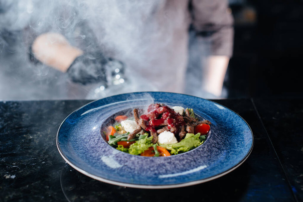 A professional chef serves a freshly prepared salad of tomato and veal greens under a glass hood with thick smoke. Beautiful smoky serving in the restaurant. - Φωτογραφία, εικόνα