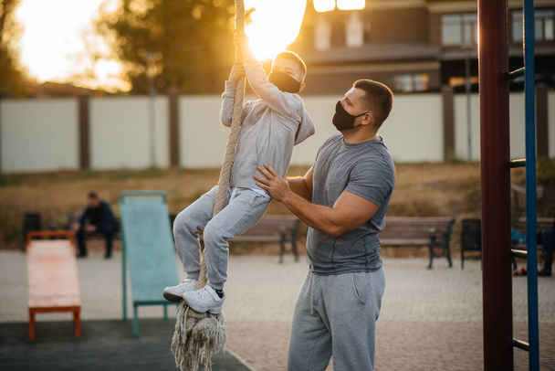 Отец помогает сыну забраться на веревку на спортивную площадку в масках во время заката. Здоровое воспитание и здоровый образ жизни - Фото, изображение