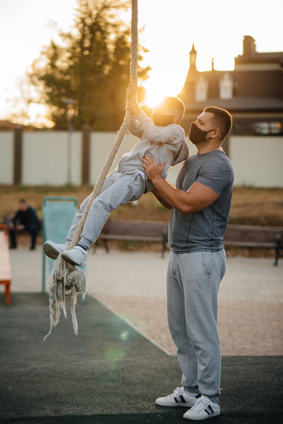 Een vader helpt zijn zoon bij het beklimmen van een touw op een sportveld met maskers tijdens zonsondergang. Gezond ouderschap en gezonde levensstijl - Foto, afbeelding