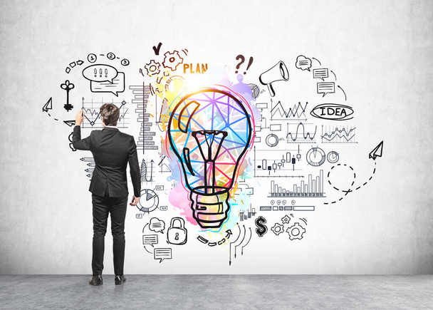 Office man in black suit, standing near colourful lightbulb drawing with network, graphs and financial analysis icons, grey office floor. Concept of new ideas, creativity and business process - Foto, imagen