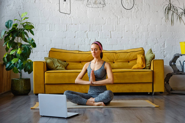 Fitness Online. Eine sportliche junge Frau streckt sich im Wohnzimmer aus, sitzt auf einer Yogamatte vor einem Laptop, kopiert den Raum. Schöne Brünette trainiert zu Hause mit Videotutorials. - Foto, Bild
